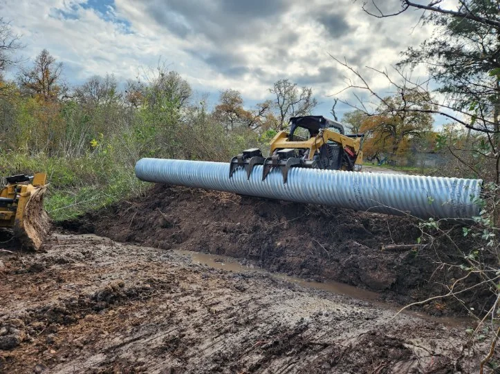 A skid steer handling a corrugated culvert pipe across an excavated drainage area.