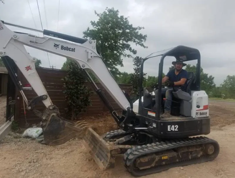 Operator in a Bobcat E42 mini excavator grading soil beside a wooden fence