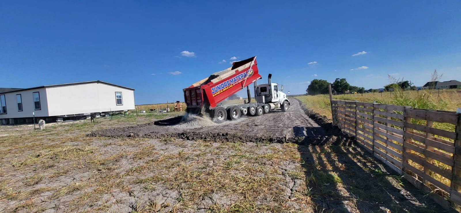 A red Juniors Materials truck dropping material beside a fence and mobile home.